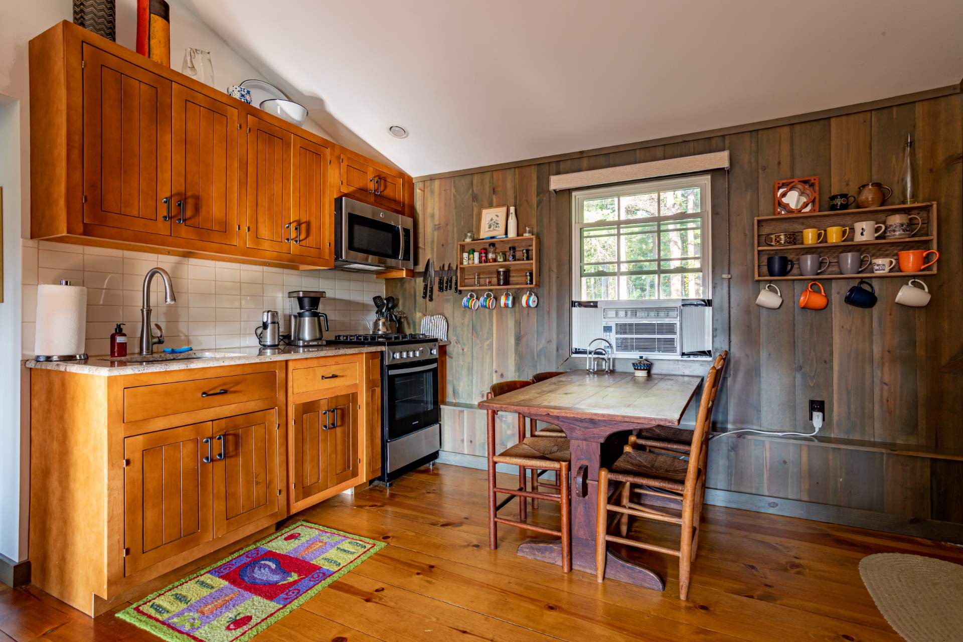 A cozy kitchen with wood-paneled walls and light wood cabinets features a stainless steel range and microwave, a white subway tile backsplash, and a rustic wooden dining table with chairs.