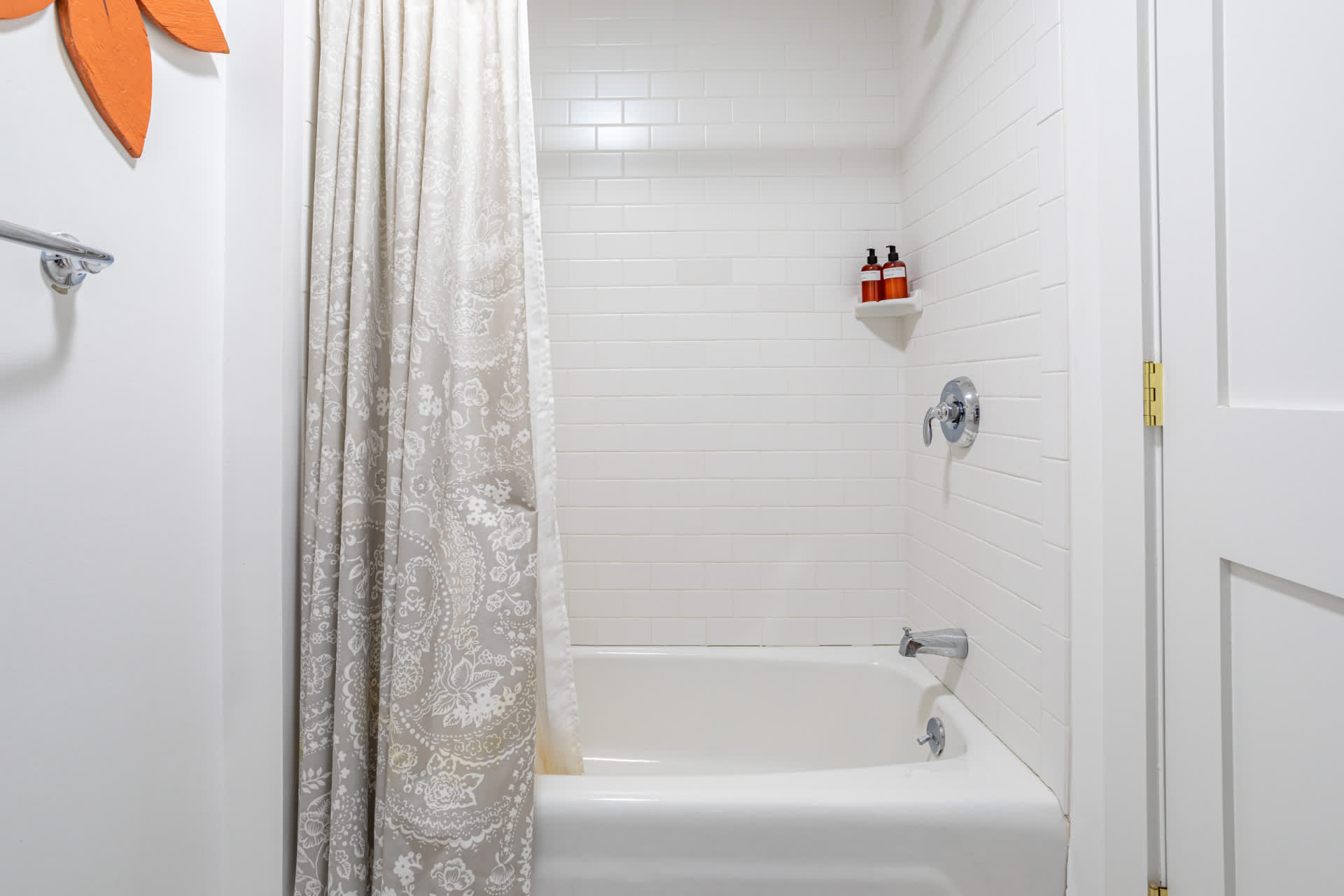 Bright bathroom featuring white subway tiled walls surrounding a clean white bathtub with a shower curtain. A small corner shelf holds toiletries, and the overall aesthetic is simple and functional.