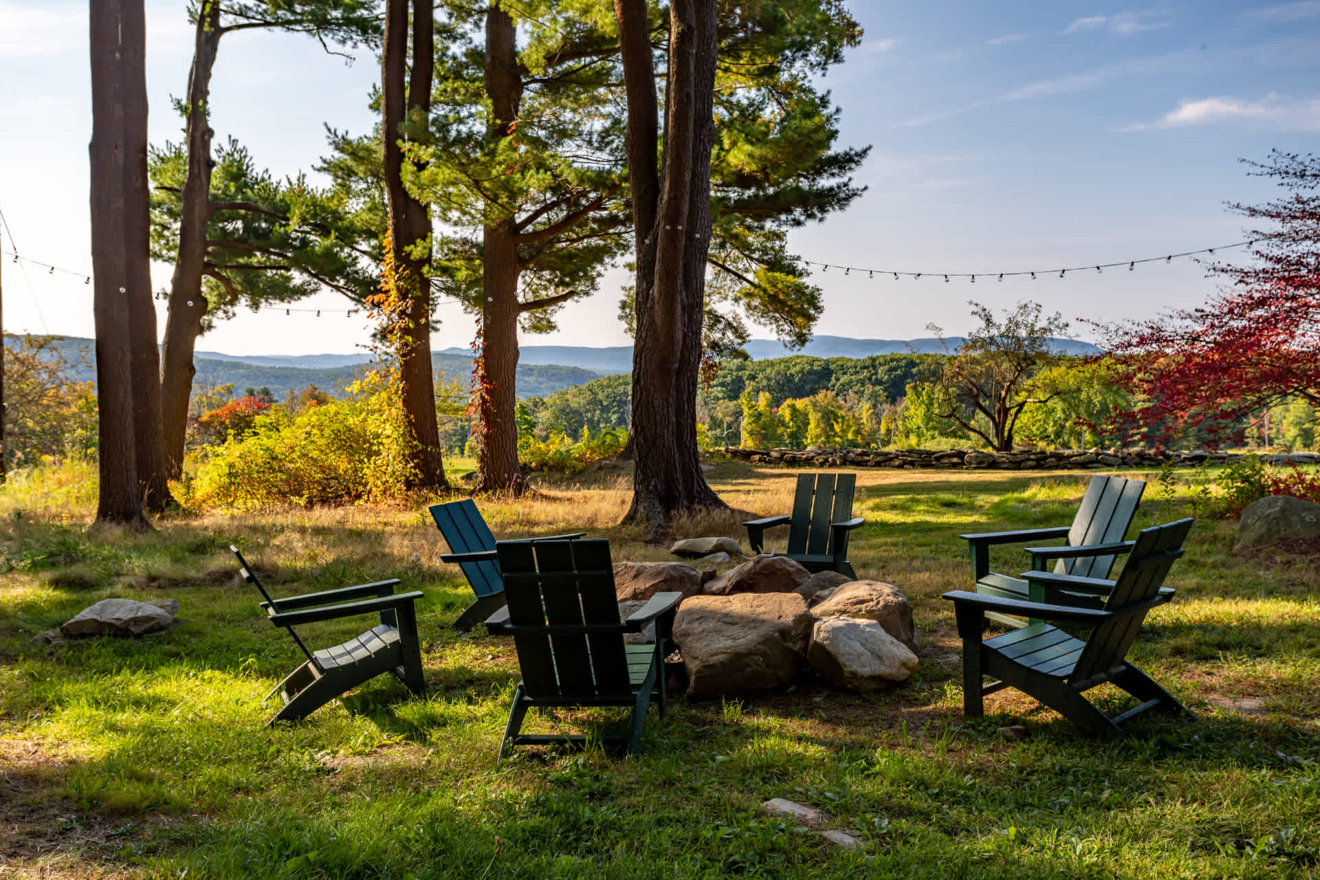 A scenic outdoor fire pit area features several green Adirondack chairs arranged around a stone fire pit, set in a grassy clearing surrounded by tall trees with distant rolling hills in the background.