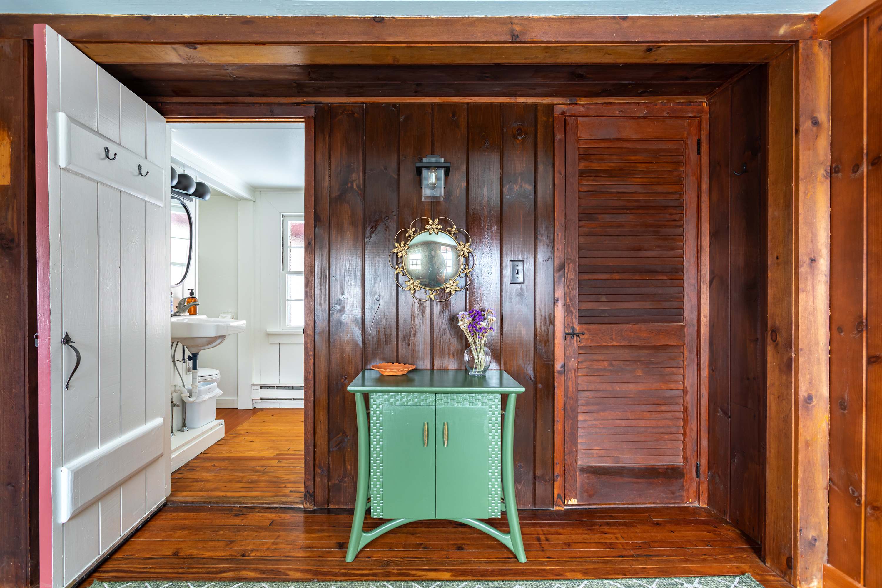 A rustic entryway with warm wooden walls and floors, featuring a small green console table with a decorative mirror and vase. A white door with hooks is open to reveal a glimpse of a bathroom with a sink, while a louvered wooden door is closed on the right.