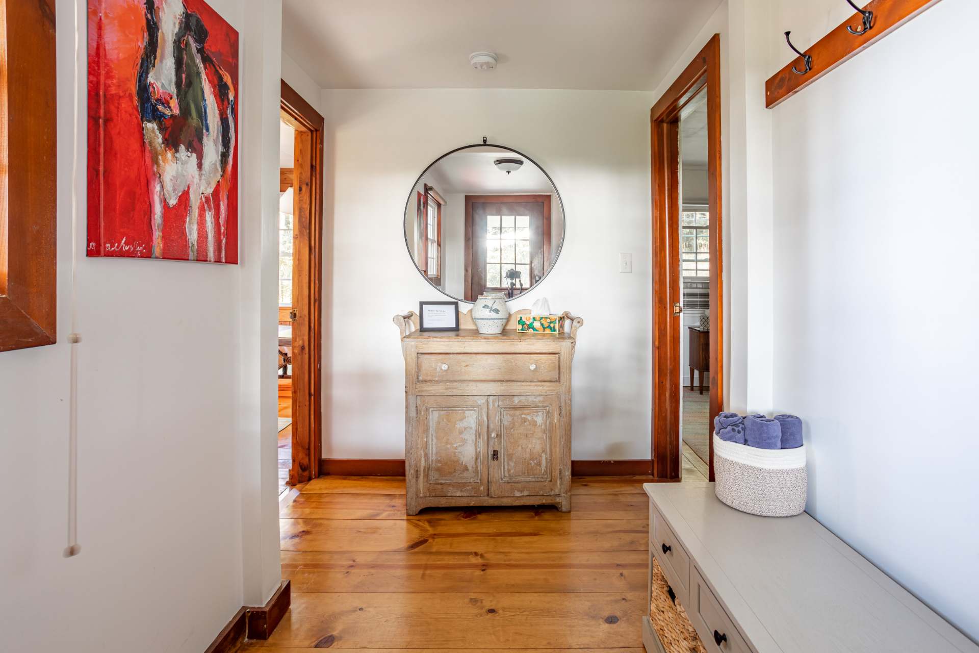 A welcoming foyer features warm hardwood floors, a rustic wooden console table with a round mirror above, and a vibrant red and white abstract painting on the left wall. Two open doorways lead to other rooms, suggesting a central hallway.