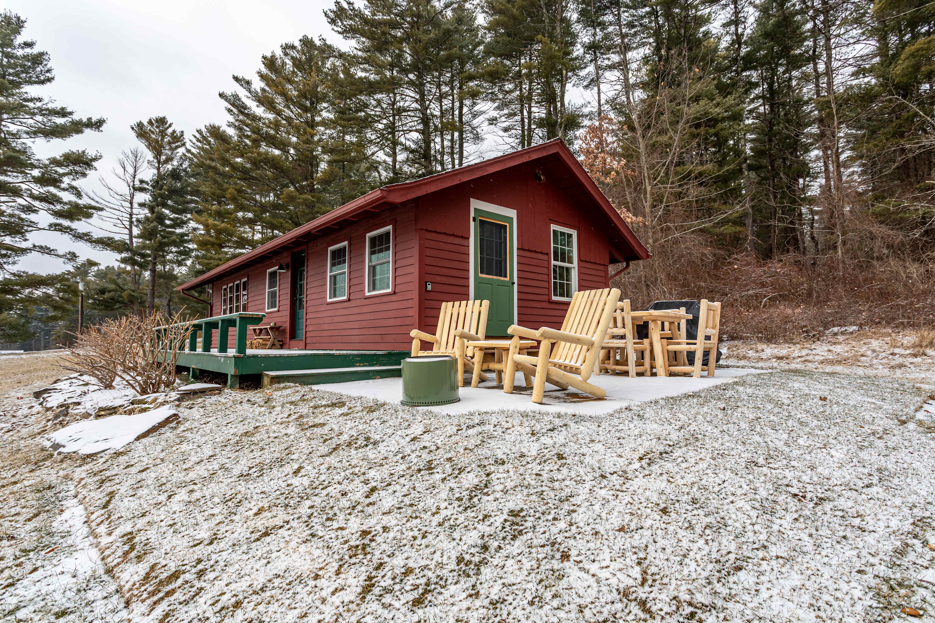 A rustic red cabin with a green door and white-trimmed windows stands in a snowy landscape, surrounded by tall pine trees. A small patio with wooden Adirondack chairs and a table is visible in the foreground.