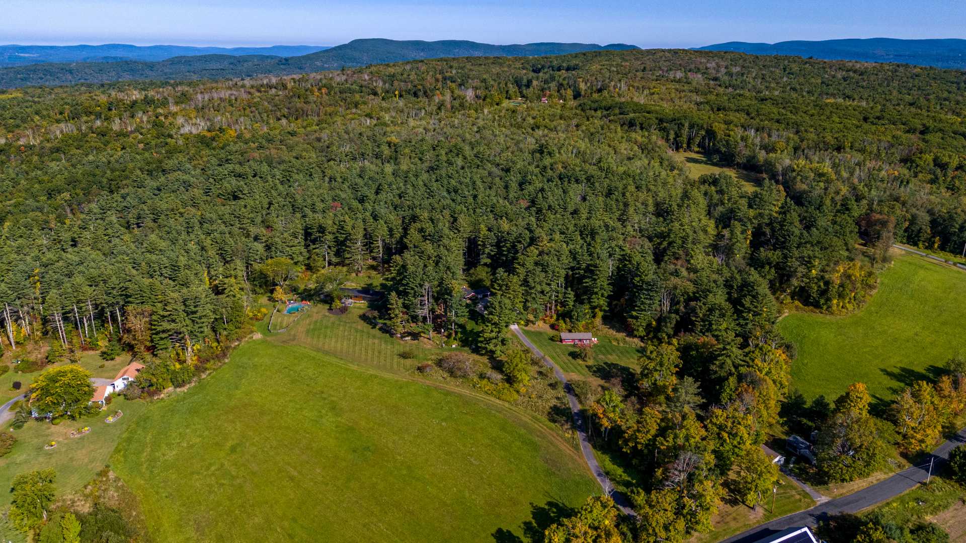 A high aerial view showcases a sprawling property with a red cottage and other structures nestled within vast green forests and open meadows, with distant rolling hills under a clear sky. A winding driveway and a swimming pool are also visible, indicating a large estate.