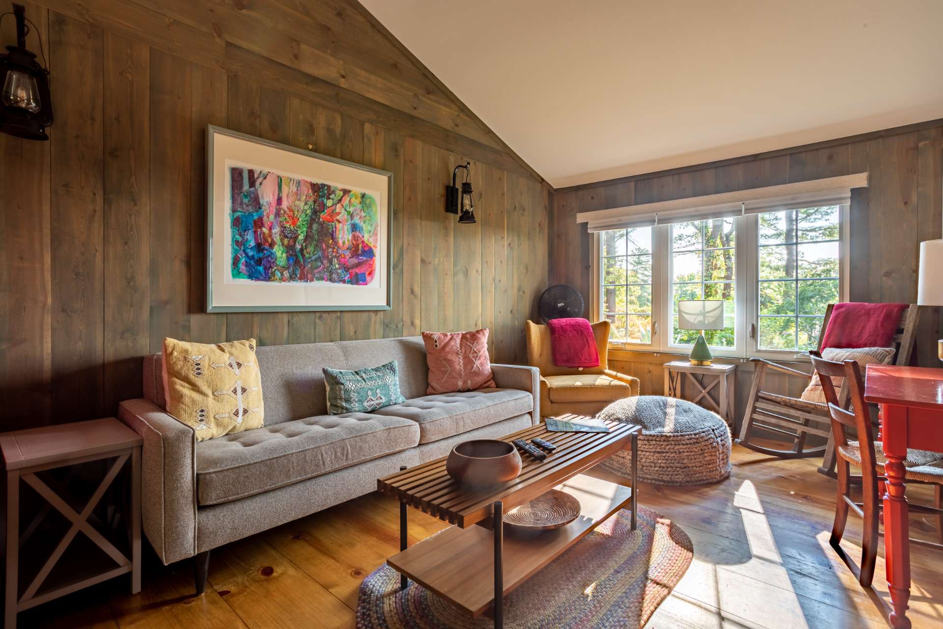 A warm and inviting living room features wood-paneled walls, a light gray sofa with colorful throw pillows, a slatted wooden coffee table, and large windows offering views of the surrounding trees.