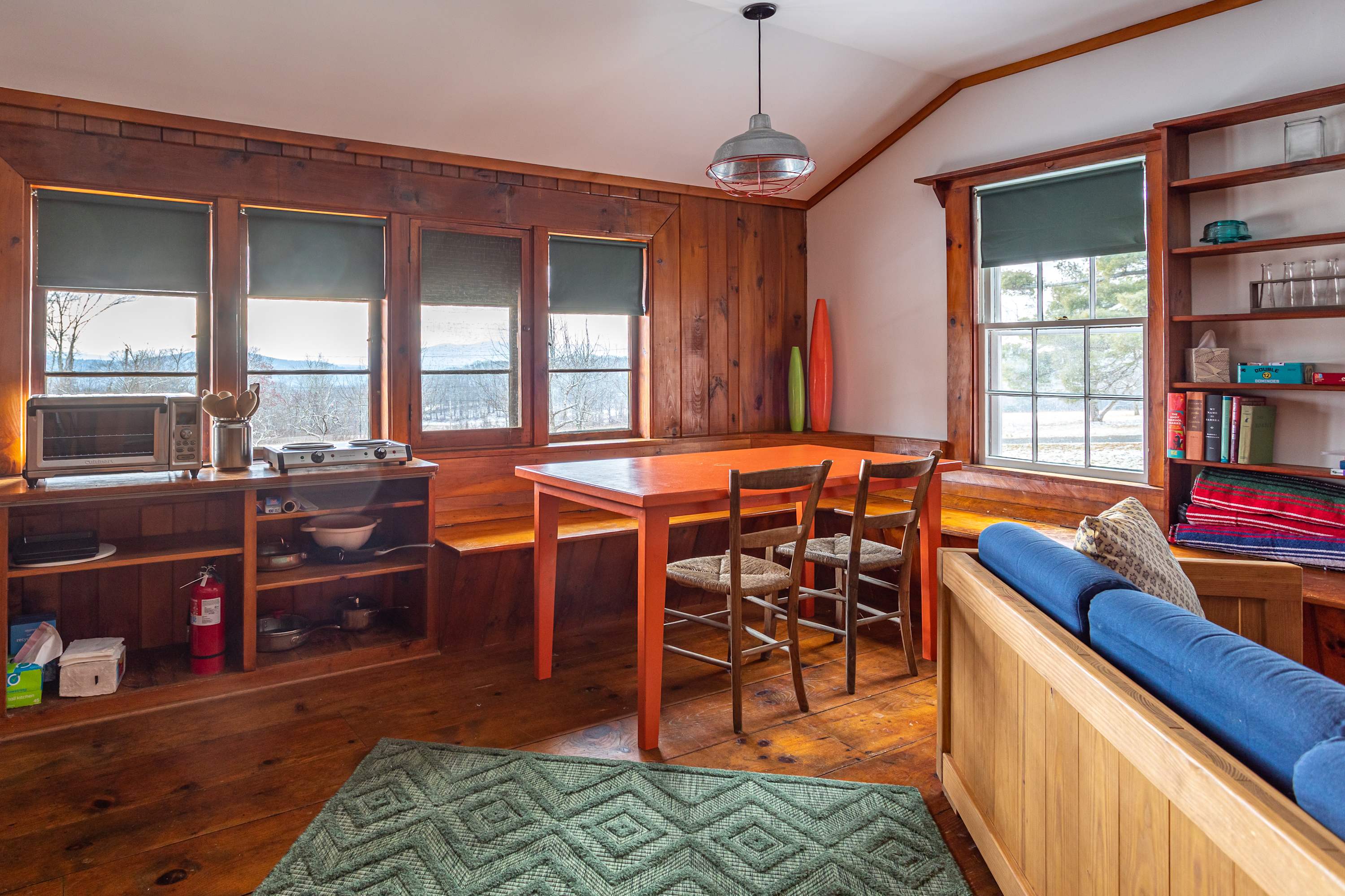A rustic dining area with warm wooden walls and floors features an orange dining table with two chairs, a built-in window bench, and a view of the snowy outdoors. A small kitchen counter with appliances is visible on the left, and a blue sofa is partially seen on the right.