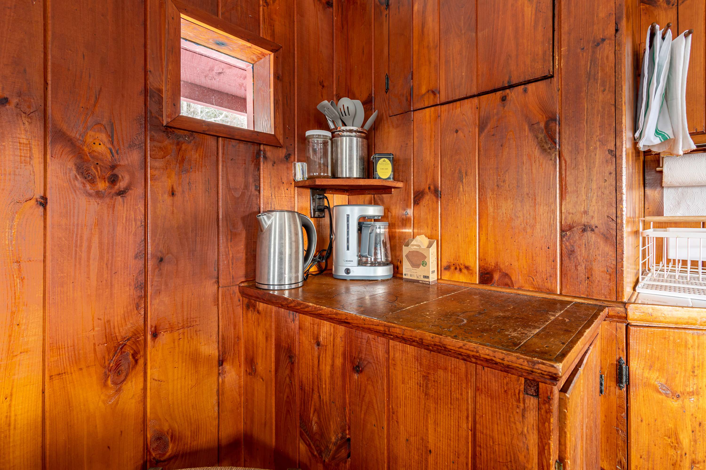 A corner of the kitchen with warm wooden paneling and a small counter space holding a silver electric kettle and a white coffee maker. A small window above the counter provides a peek outside, and kitchen utensils are stored in a jar.