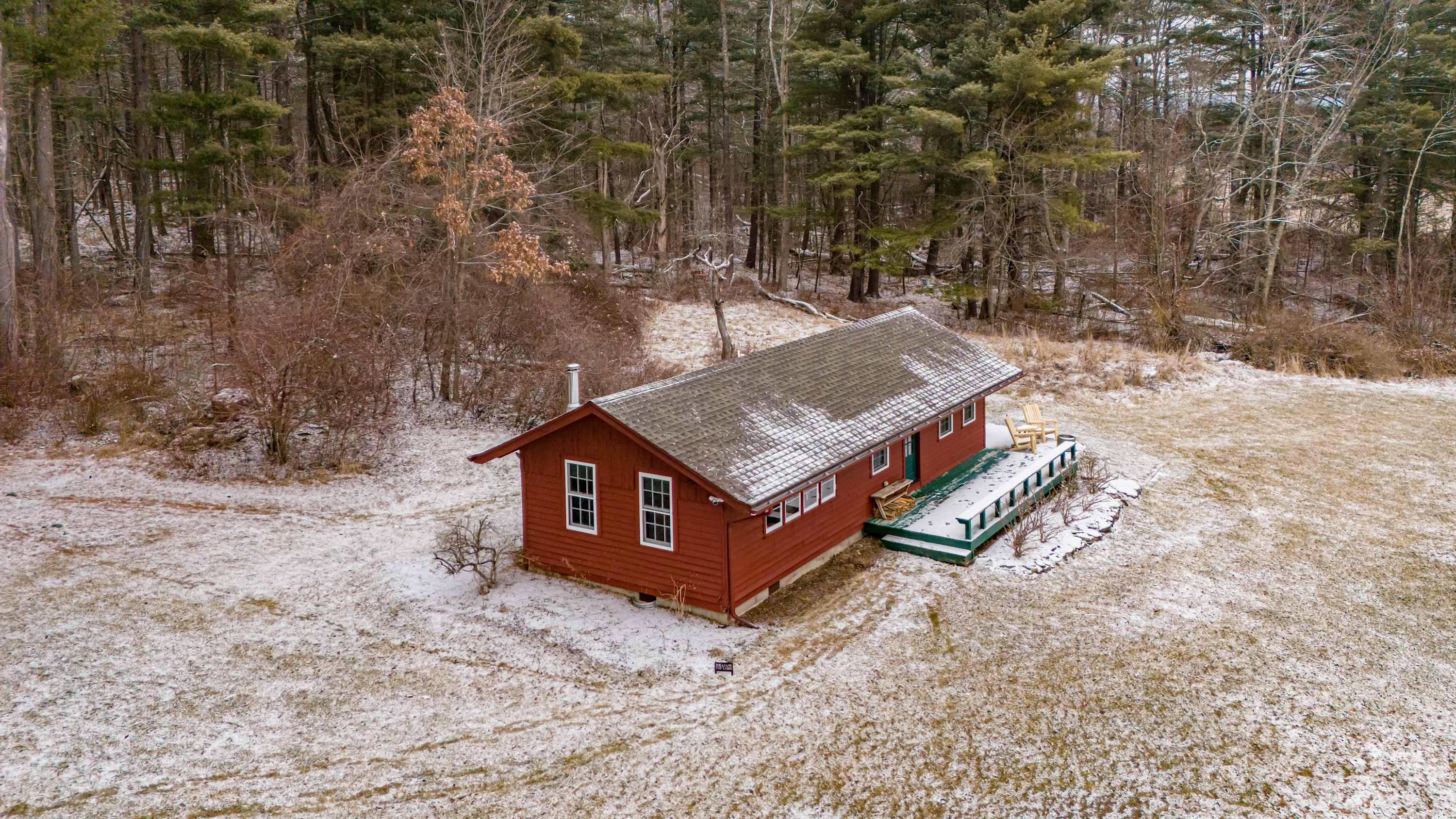 An aerial view of a long, red single-story cottage with a dark roof, surrounded by a light dusting of snow on a grassy field, backed by a dense, bare winter forest. A green and white deck with outdoor seating is visible on the right side of the cottage.