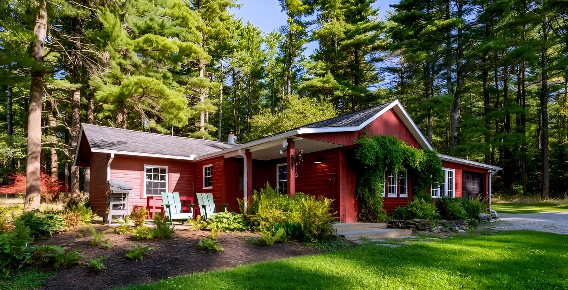 A charming red cottage with white trim is nestled among tall green trees, featuring a covered porch and a small patio area with Adirondack chairs and a grill. Lush green grass and landscaping surround the home.