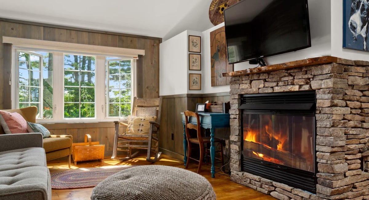 A sunlit living room showcases wood-paneled walls, a comfortable sofa, a rustic wooden rocking chair, and a prominent stone fireplace with a roaring fire and a TV mounted above. Large windows offer views of the outdoors.