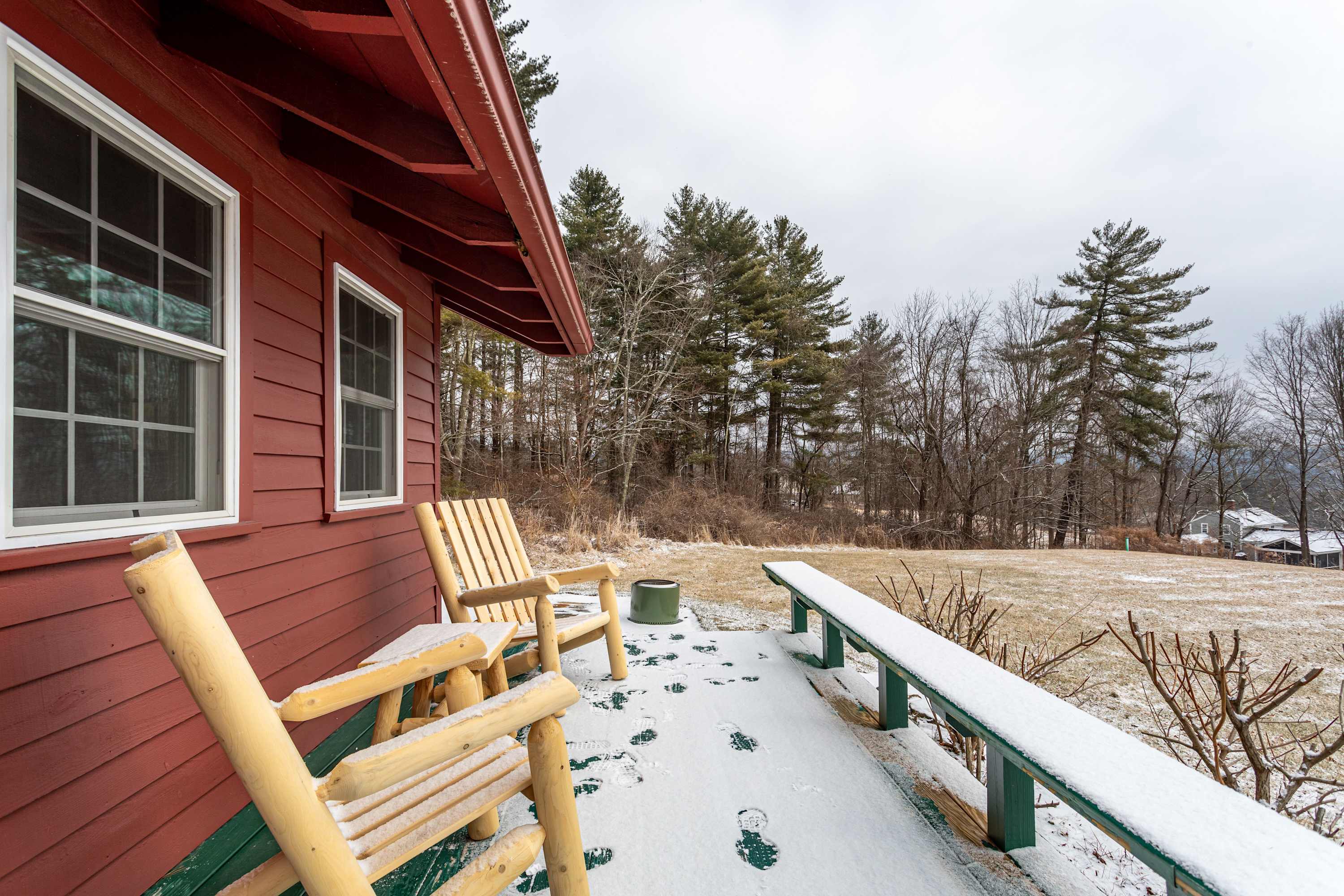 A snowy outdoor porch with a red-sided house and green trim, featuring two rustic wooden Adirondack chairs and a long wooden bench. The porch overlooks a snow-dusted field and a forest of pine trees under a cloudy sky.