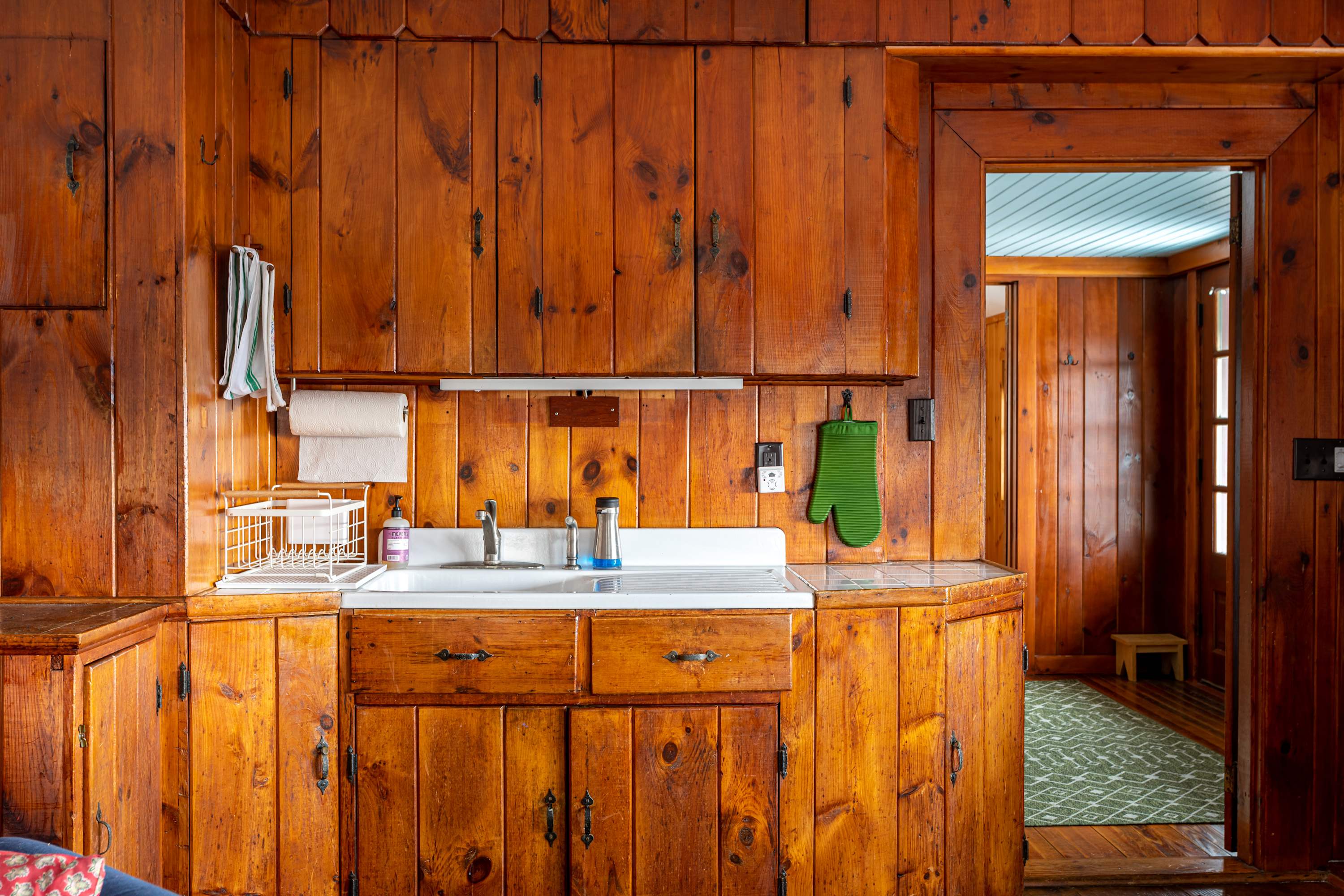 A close-up of the kitchen area, highlighting the warm wooden cabinets and a white double sink with a faucet. Paper towels hang conveniently, and a green oven mitt adds a pop of color, with a glimpse of a doorway to another room.