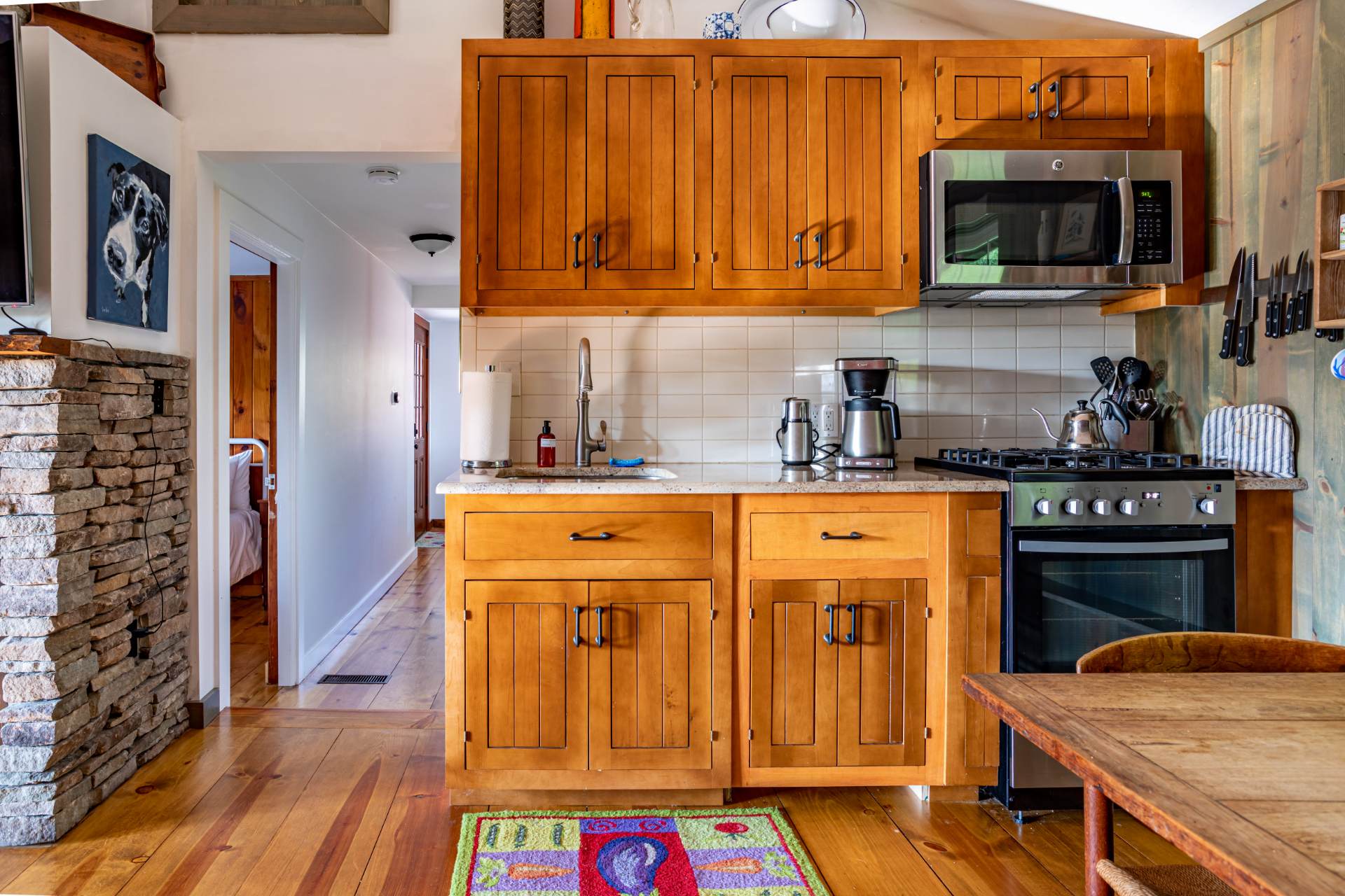 A bright kitchen features light wood cabinetry, granite countertops, a stainless steel sink, and a gas range, with a colorful rug on the hardwood floor. A stone fireplace is visible in the adjacent room.