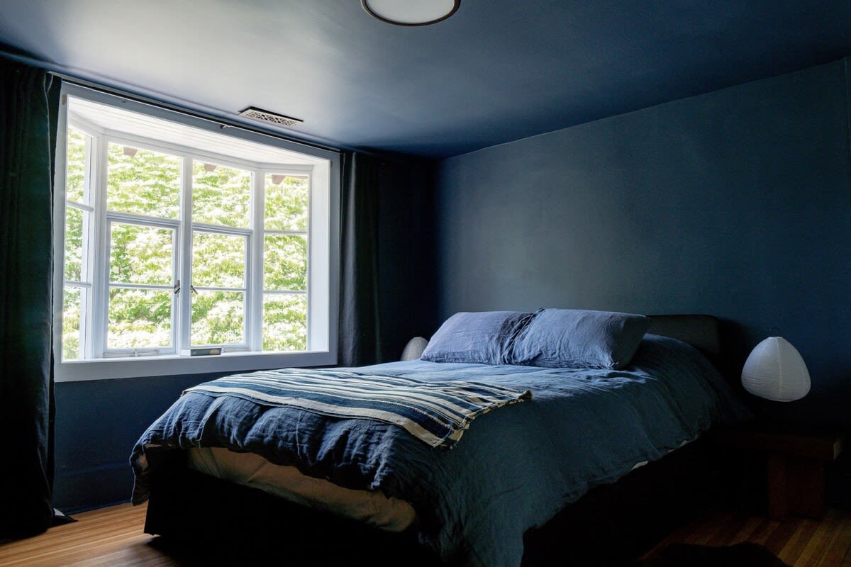 A serene bedroom with dark blue walls and ceiling, featuring a large bed with blue linens and pillows, a bay window, and a modern bedside lamp.