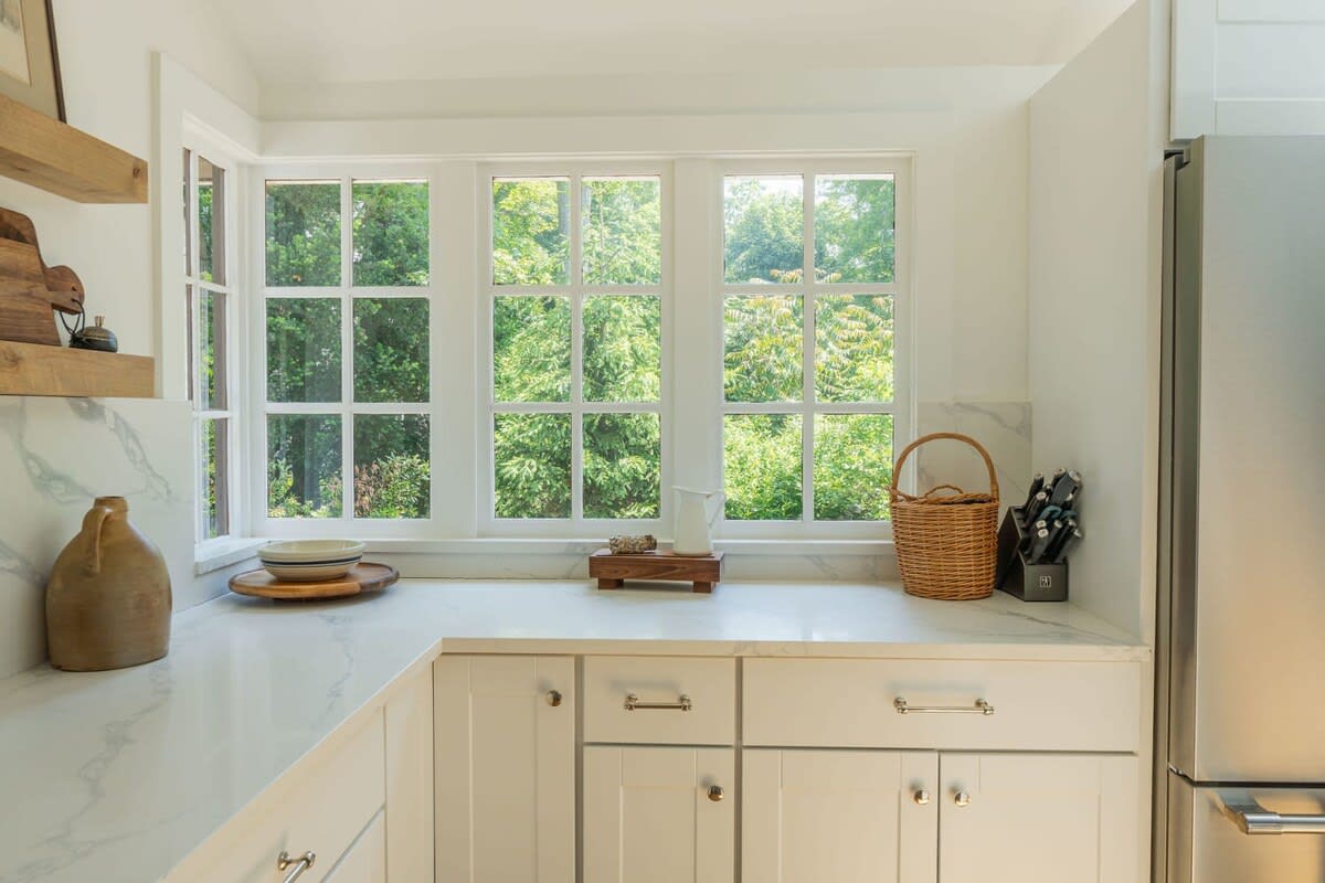 A bright kitchen corner with white cabinets and light countertops, featuring a large window with multiple panes offering views of lush outdoor greenery, and open wooden shelves.