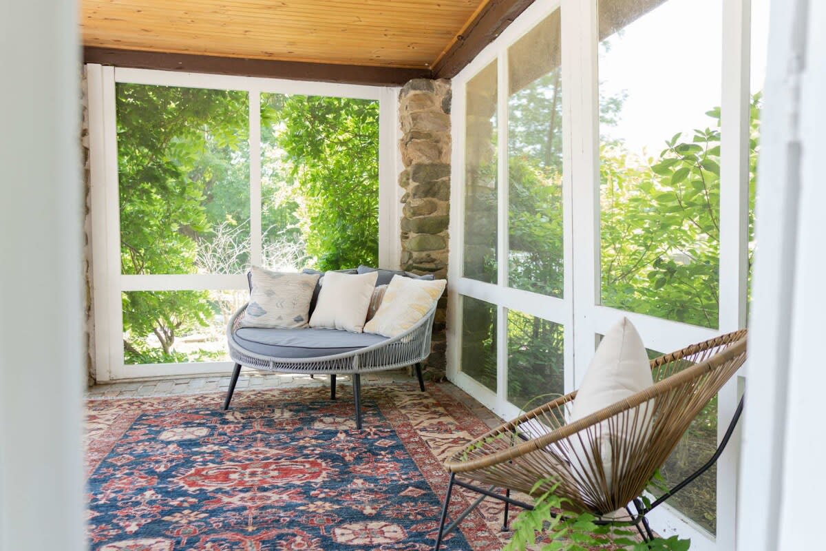 Enclosed screened porch with a cozy curved sofa and a wicker armchair, adorned with a colorful patterned rug, offering views of lush outdoor greenery through large windows.
