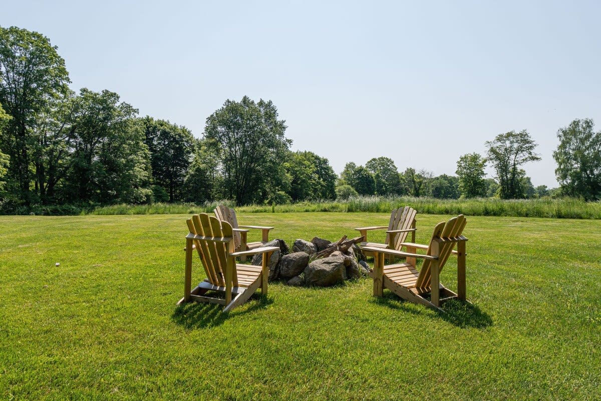 Outdoor fire pit area in a spacious green lawn, featuring a stone fire pit surrounded by four wooden Adirondack chairs, with a backdrop of trees and an open field under a clear sky.