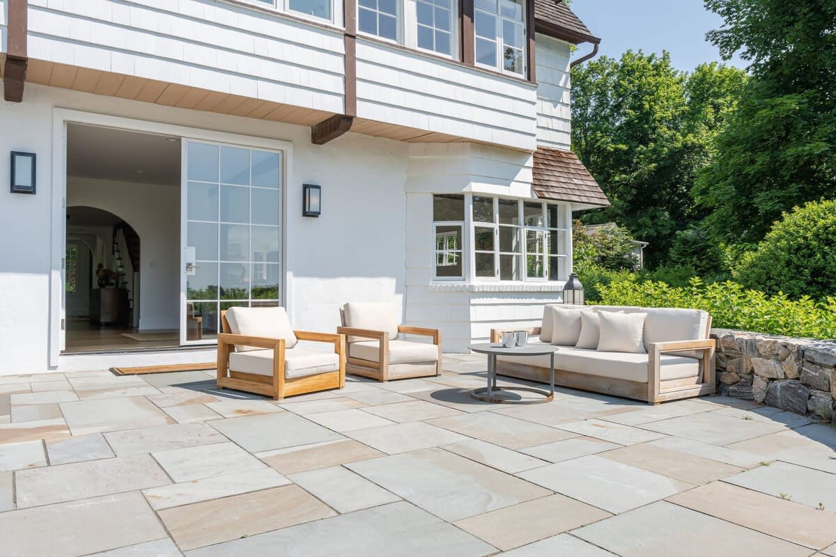 An expansive stone patio extends from the house, furnished with a modern outdoor sofa and two armchairs with light cushions. A low stone wall borders the patio, with lush greenery and trees in the background.