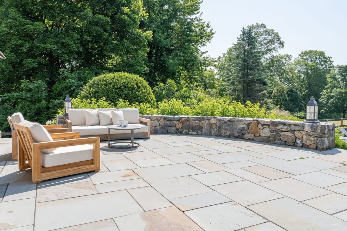 A wide shot of the stone patio with modern outdoor furniture, including a sofa and armchairs, set against a backdrop of a low stone wall and dense green foliage. The sunny outdoor space offers a serene view of the surrounding nature.