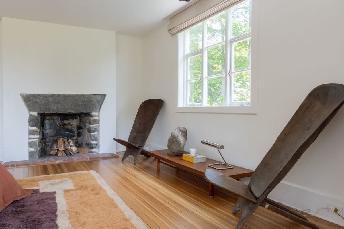 Cozy living area with a rustic stone fireplace, two distinctive wooden lounge chairs, and a large window overlooking lush greenery. Hardwood floors are partially covered by a patterned rug.