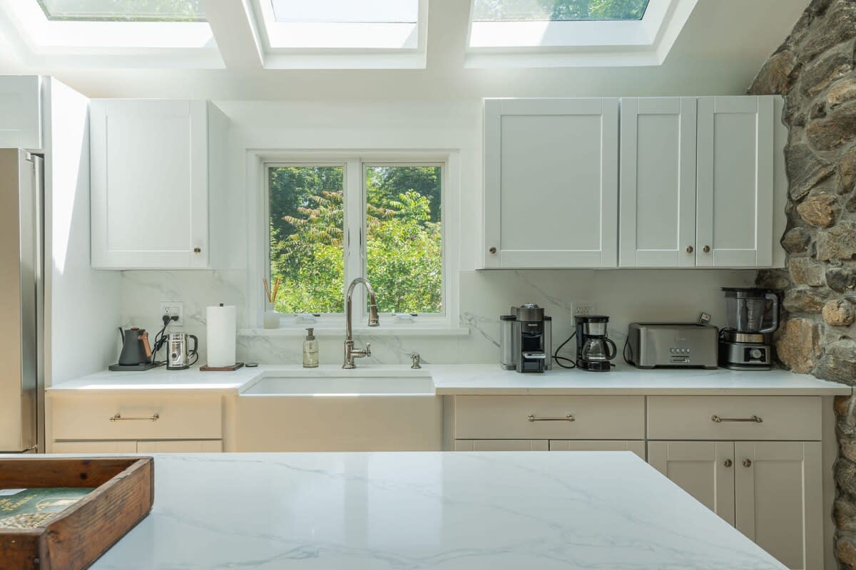A well-lit kitchen counter area with a farmhouse sink, white cabinets, and light countertops, featuring a window overlooking trees and several small appliances.