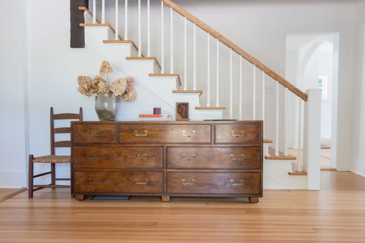 A welcoming foyer with light hardwood floors and a white staircase with a wooden handrail, featuring a large antique wooden dresser adorned with dried flowers and decor.