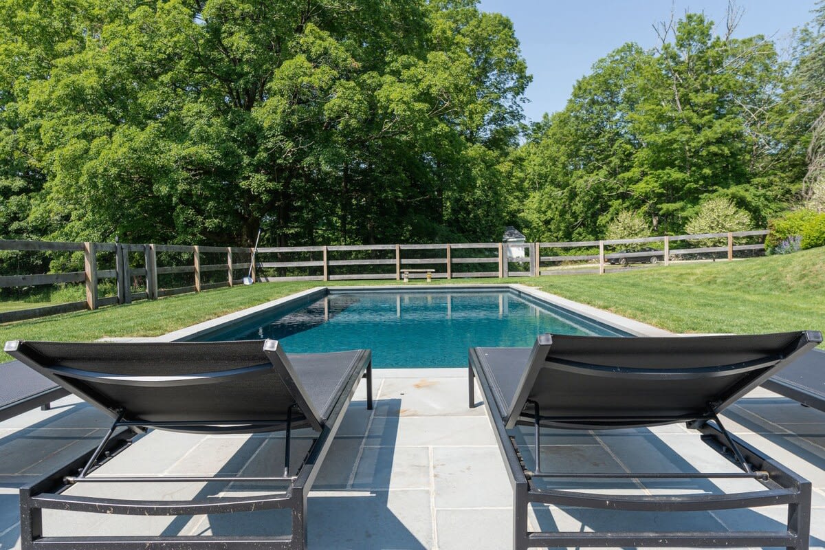 Rectangular inground swimming pool with dark blue water, surrounded by a light grey stone patio and green grass. Two black modern lounge chairs are in the foreground, with a wooden fence and lush green trees in the background.