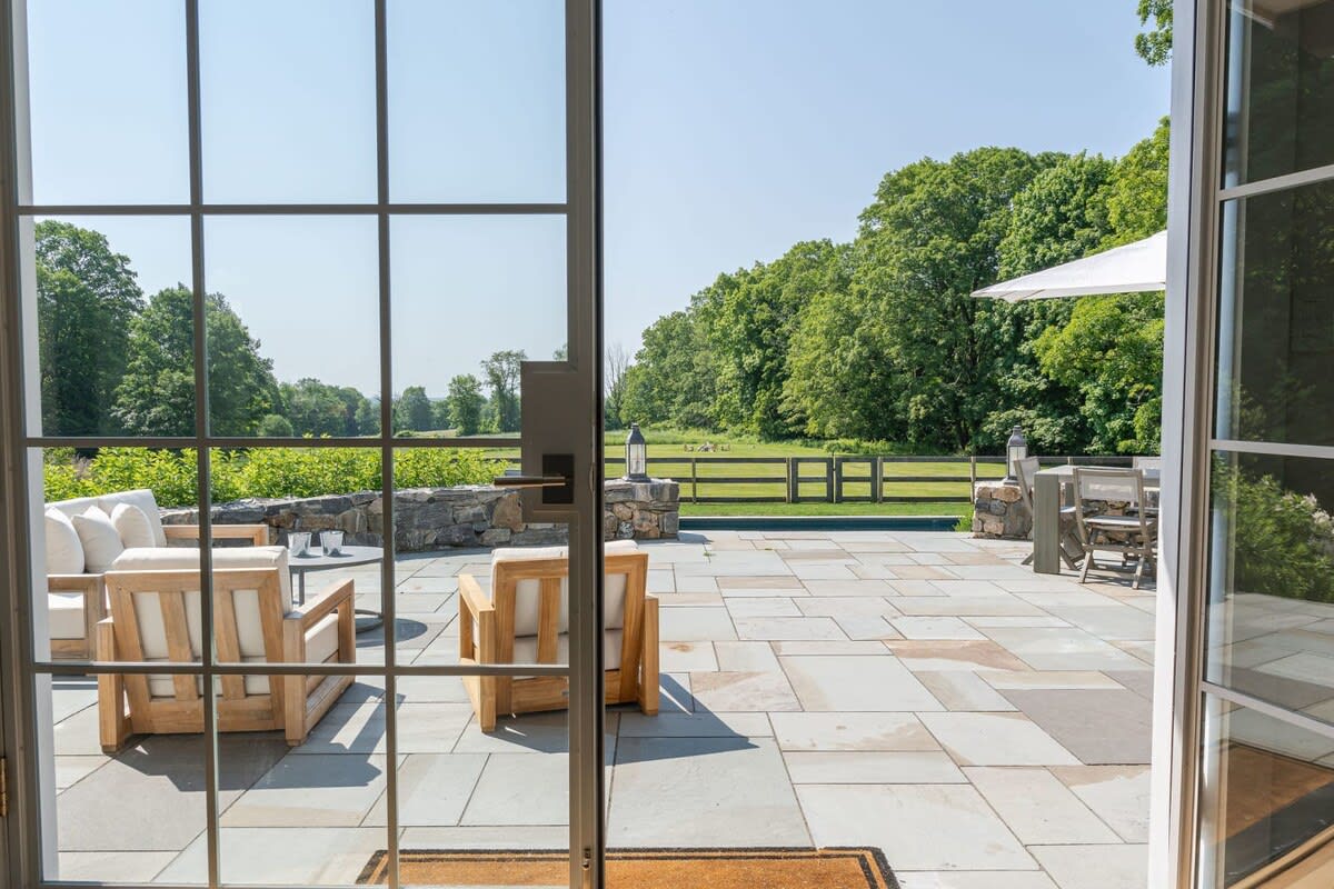View from inside looking out onto a large stone patio with modern outdoor seating, including a sofa and armchairs with light cushions. Beyond the patio, a lush green landscape with trees and a distant pool is visible.