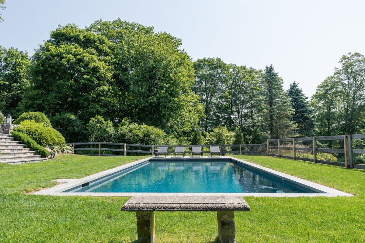 Wide shot of a rectangular inground swimming pool with dark blue water, set in a manicured green lawn. A stone bench sits in the foreground, and four black lounge chairs are visible at the far end of the pool. A wooden fence and dense green trees line the property.