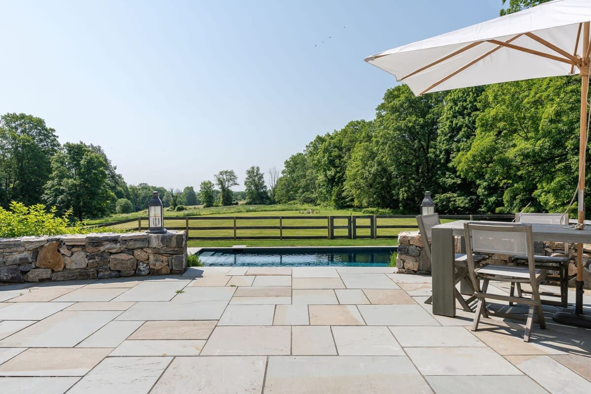 A spacious stone patio features an outdoor dining table with an umbrella, overlooking a rectangular swimming pool and a vast green field bordered by a wooden fence and trees. Decorative lanterns are placed on the stone wall.