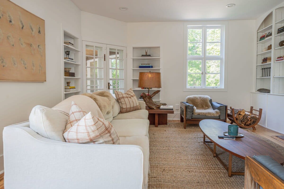 A cozy living room with a white sofa, a dark wood coffee table, and a jute rug, featuring built-in shelves, a large window, and a rustic log holder next to an armchair.