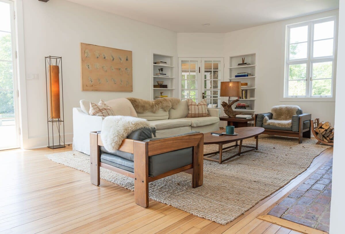 Bright living room with a cream sofa and two wooden armchairs, all adorned with faux fur throws, centered around a wooden coffee table on a large jute rug, with built-in shelves and French doors.