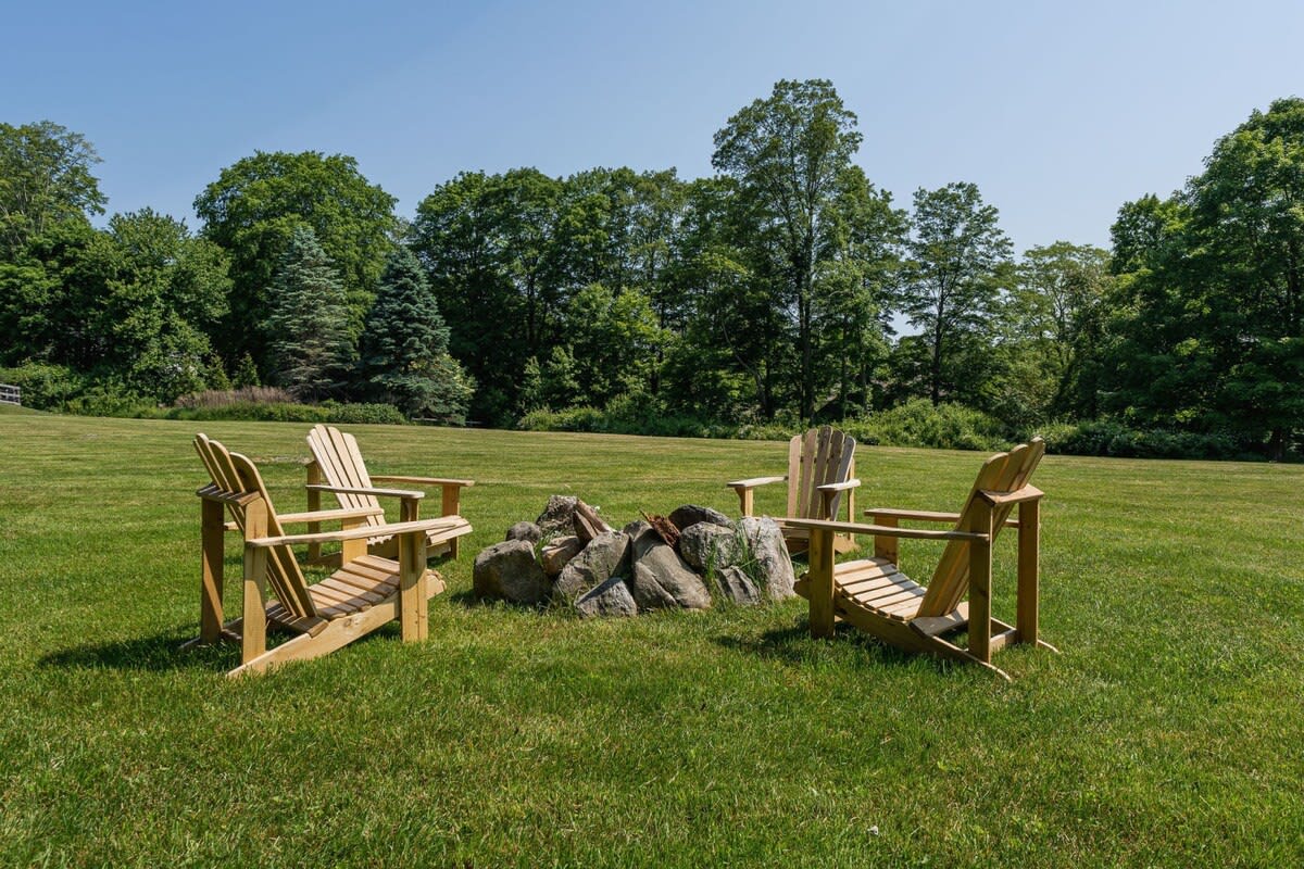 A rustic fire pit area is set in a large grassy field, surrounded by four wooden Adirondack chairs. The fire pit is constructed from natural stones, with dense green trees forming the backdrop.