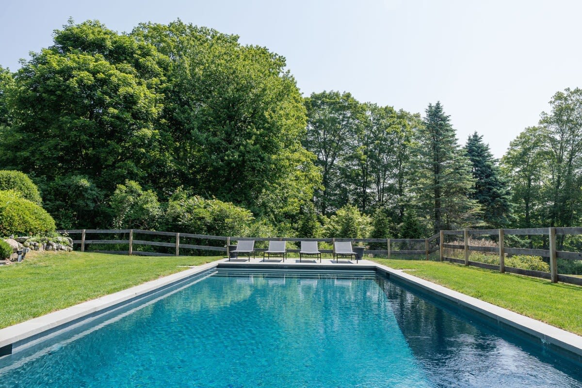 View of the rectangular inground swimming pool from the opposite end, showing four lounge chairs, surrounded by lush green grass and a wooden fence, with a dense forest line in the background.