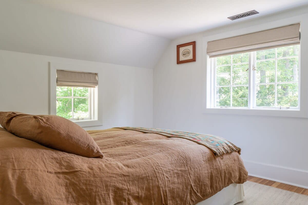 Bright bedroom with white walls and two windows fitted with room-darkening shades, offering privacy and light control. A bed with rust-colored linen bedding is visible, along with hardwood floors.