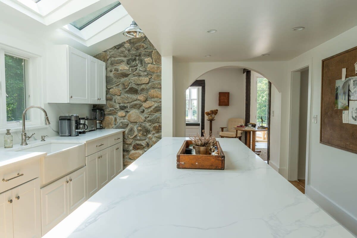 A spacious kitchen featuring a large white island with a farmhouse sink, white cabinets, and a prominent stone accent wall, illuminated by skylights and leading to another room through an archway.
