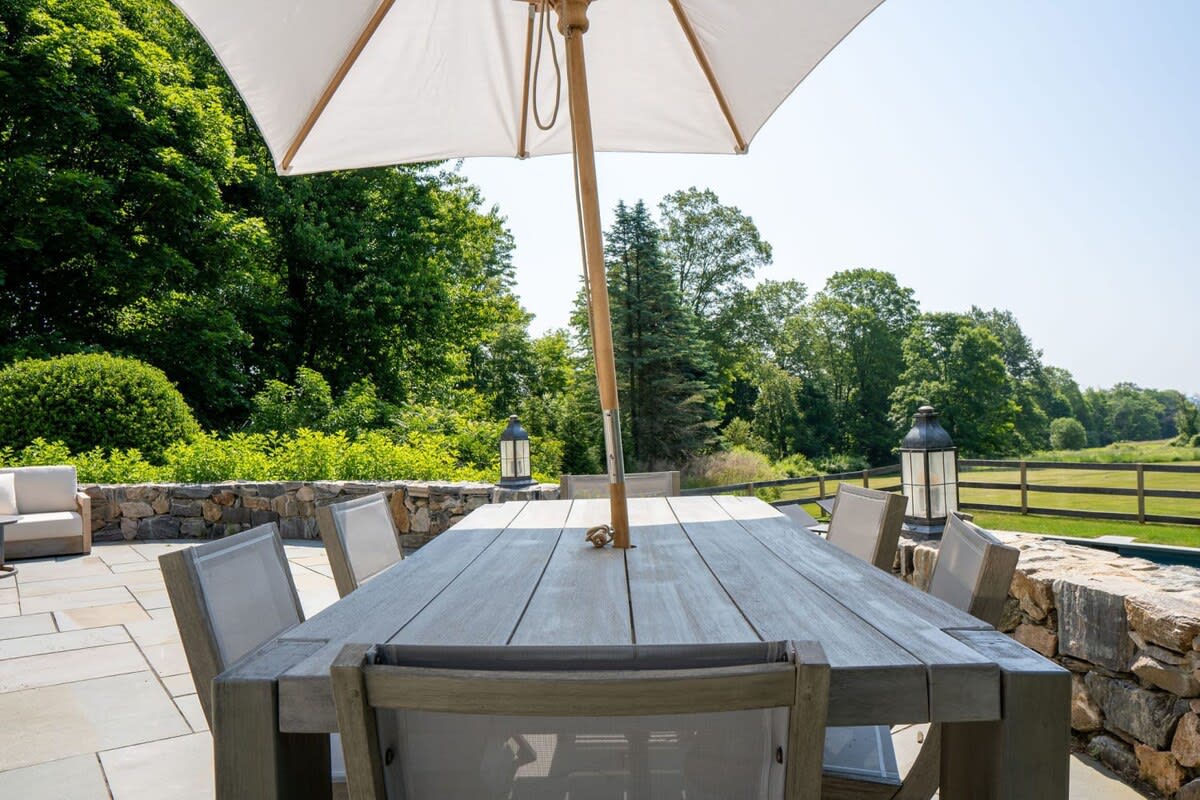 Close-up of a large wooden outdoor dining table with an umbrella, set on a stone patio. The background shows lush green trees and a glimpse of the pool area, creating an inviting al fresco dining setting.