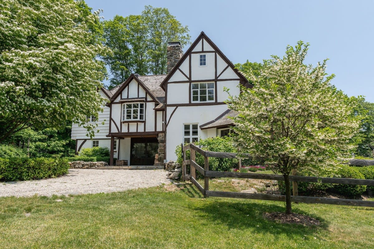 The front exterior of a charming white Tudor-style house with dark timber framing and a shingled roof, surrounded by lush green trees and a well-maintained lawn. A gravel driveway is visible in the foreground, bordered by a rustic wooden fence and a small flowering tree.