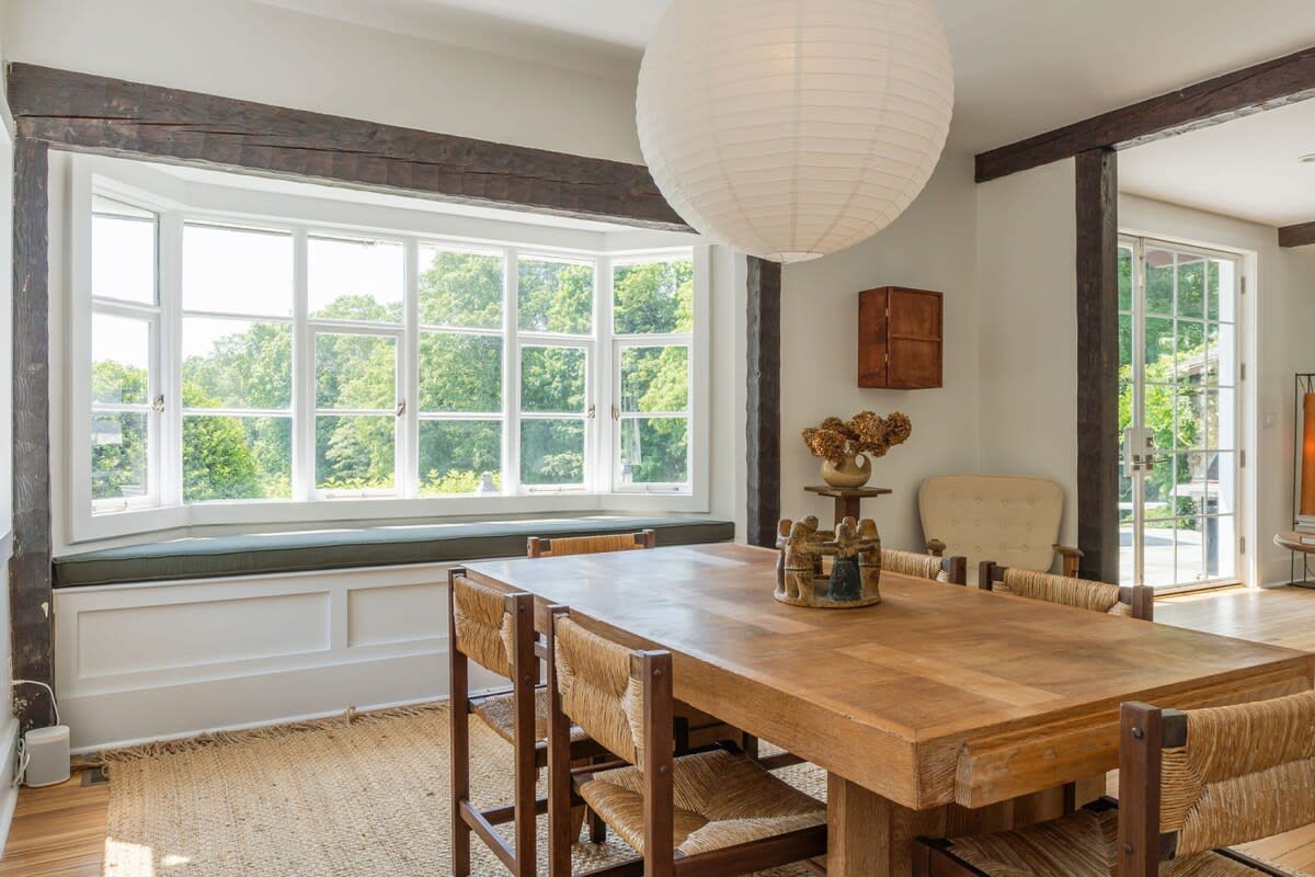 Rustic dining room with a large wooden dining table and woven chairs, a prominent bay window with a built-in bench overlooking greenery, and a large white paper lantern light fixture.