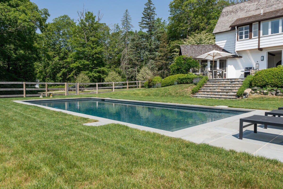 A rectangular swimming pool with a dark liner is surrounded by a stone patio and a well-maintained green lawn. The house, with its white siding and brown trim, is visible on a slight incline in the background, along with mature trees.
