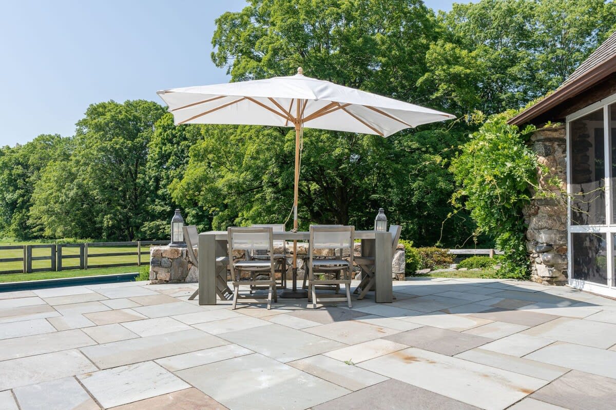 Outdoor patio area with large flagstone pavers, featuring a grey outdoor dining table with chairs and a large white umbrella, adjacent to a stone wall and lush greenery.