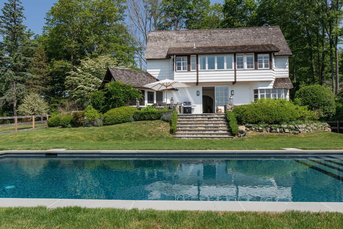 View of the white two-story house with dark trim and a shingled roof, seen from across the inground swimming pool. A stone patio with steps leads up to a back patio area with an umbrella. The pool's dark blue water reflects the sky, and the property is surrounded by green lawns and mature trees.