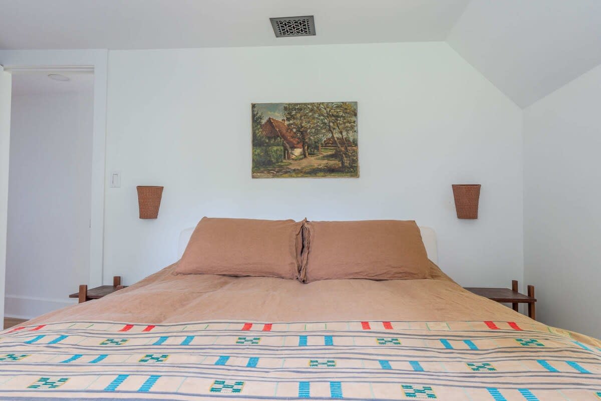A cozy bedroom features a bed with brown linen bedding and a patterned blanket, flanked by two small wooden nightstands and woven wall sconces. A landscape painting hangs above the bed on the white wall.