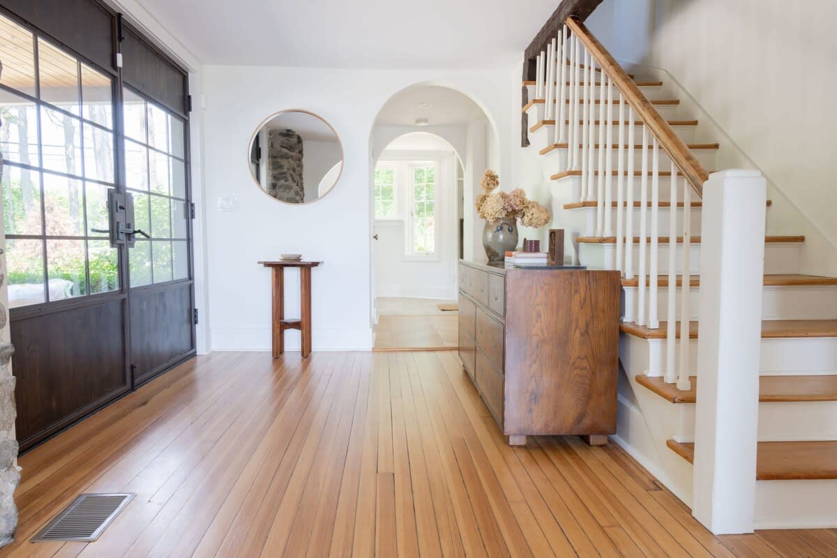 A spacious foyer with warm hardwood floors and white walls features a dark-framed glass entry door and a wooden staircase with white balusters. A round mirror hangs above a small wooden table, and a wooden dresser is placed near the stairs.