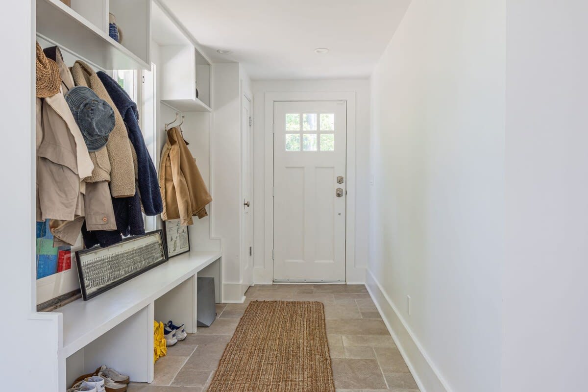 Bright mudroom featuring a built-in white storage bench, coat hooks with various jackets, and a white front door with glass panes. A woven rug covers the light tiled floor.