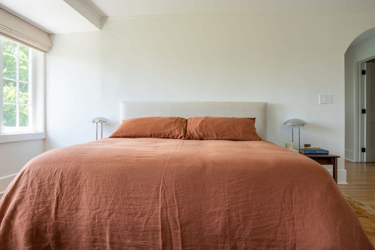 Bright bedroom featuring a queen-sized bed with a light upholstered headboard and rust-colored linen bedding. Two modern lamps sit on wooden nightstands, complementing the warm tones.