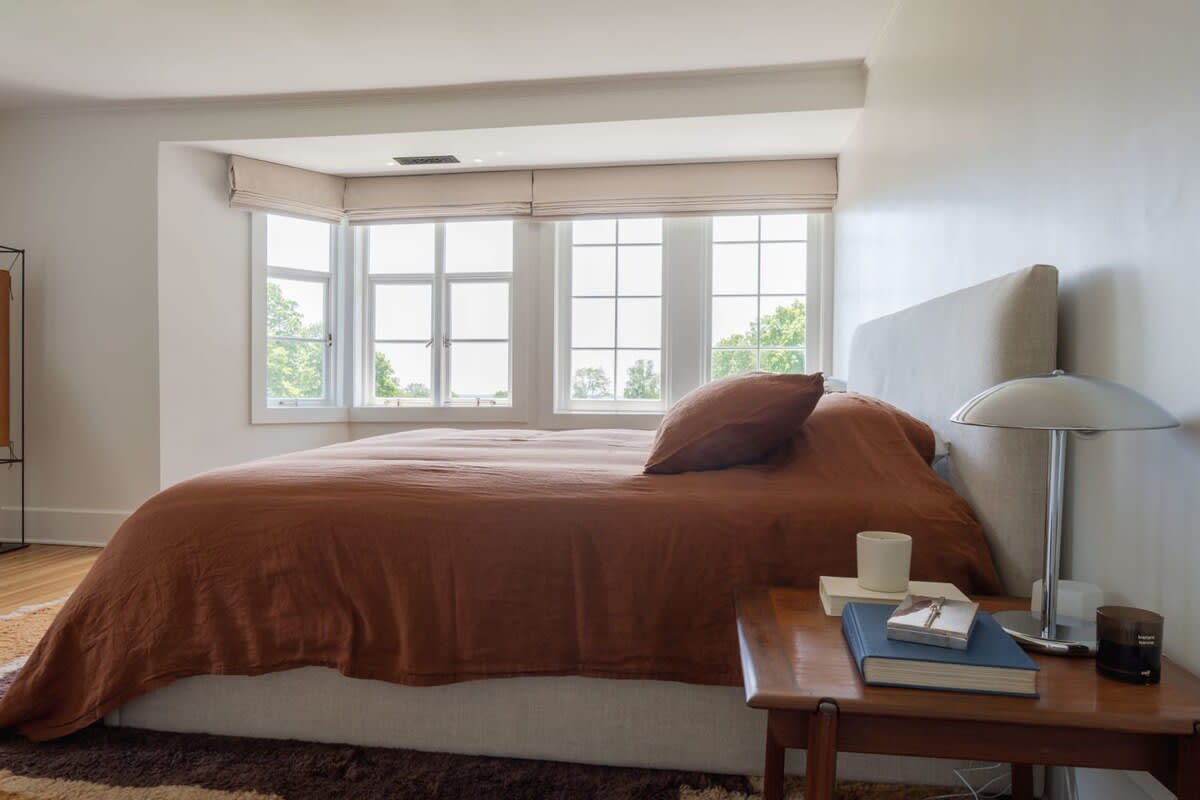 Spacious bedroom with a queen-sized bed featuring a light upholstered headboard and rust-colored linen bedding. Large bay windows offer views of lush greenery, filling the room with natural light.