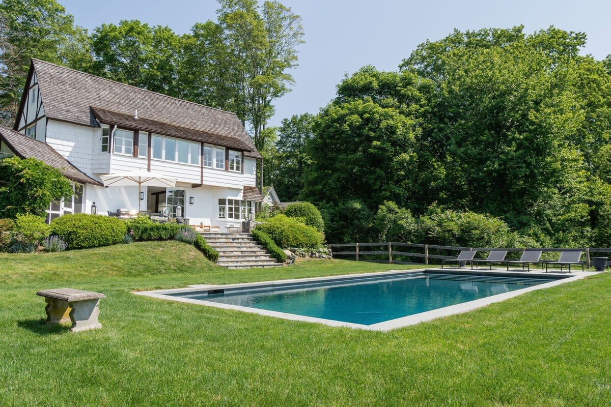 The back of a white two-story house with dark trim and a shingled roof, featuring multiple windows and a patio area with an umbrella. A stone staircase leads down to a large green lawn with a rectangular inground pool, surrounded by lush trees and a wooden fence.