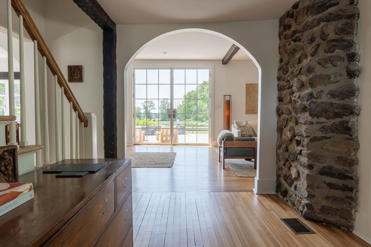 A bright foyer with light hardwood floors and white walls, featuring a rustic stone column on the right and an arched doorway leading to a living space with large windows overlooking greenery.