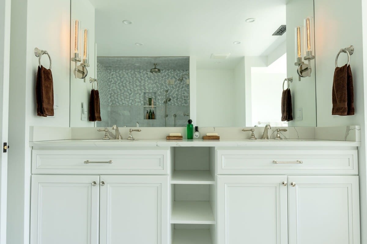 Elegant double vanity with white shaker cabinets, a white marble countertop, and two sinks. A large mirror reflects the shower, flanked by modern sconce lighting fixtures.