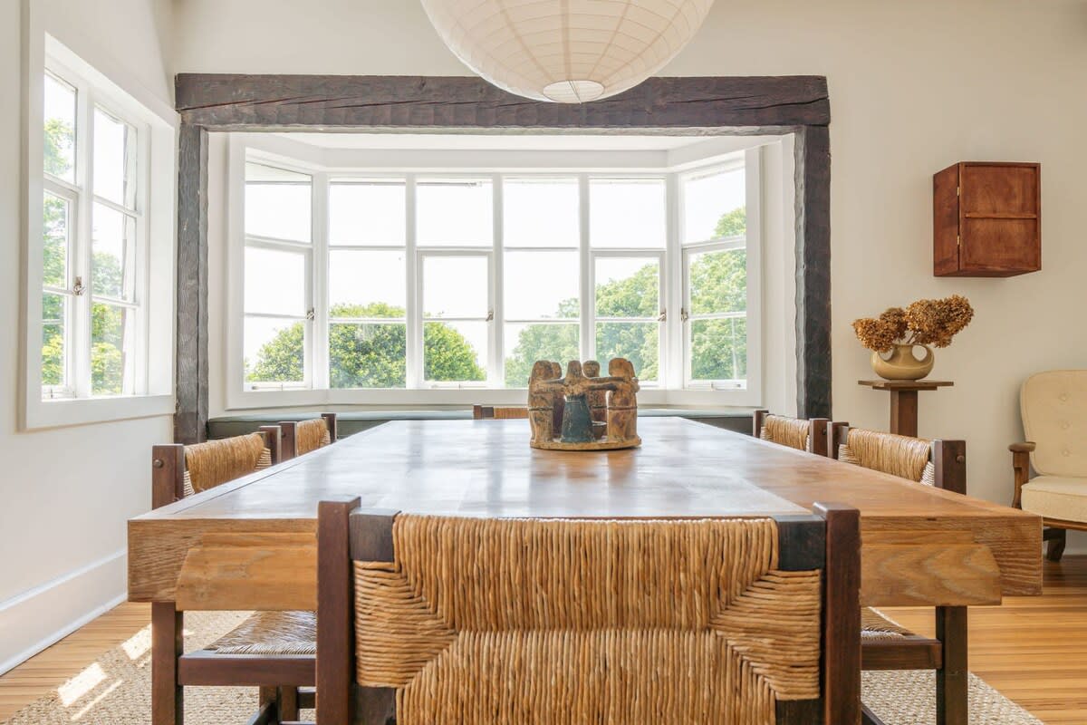 A rustic dining room featuring a large wooden table with woven-back chairs, set against a bay window offering expansive views of lush greenery, with exposed wooden beams overhead.
